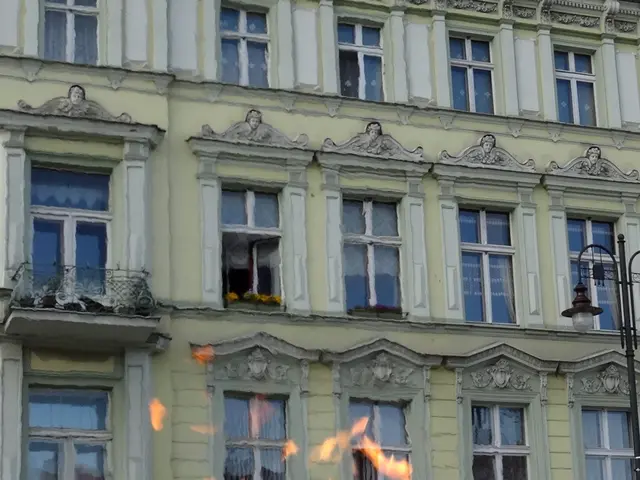 The image shows a large fire burning in front of a building with windows and doors, a light pole,...