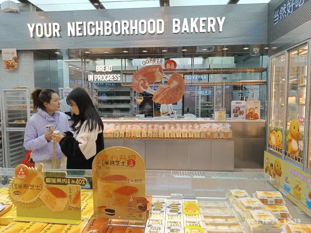 The image shows two women standing in front of a bakery counter, looking at their cell phones. On...
