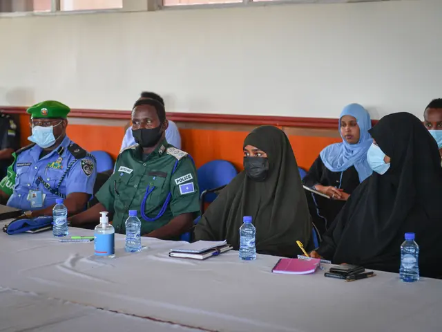 The image shows a group of people sitting around a long table with water bottles, books, pens,...