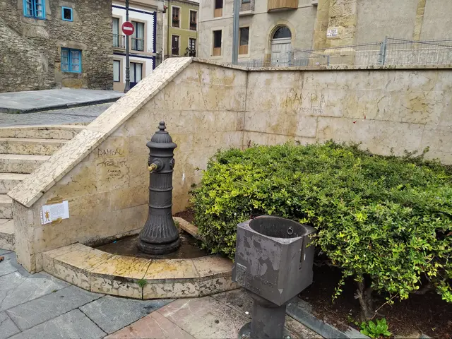 The image shows a small fountain in the middle of a city square surrounded by buildings with...