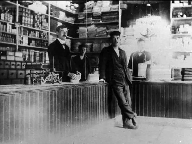 The image shows a black and white photo of three men standing at a counter in a store. On the...