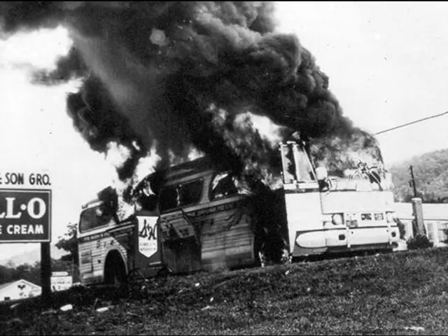 The image shows a black and white photo of a bus engulfed in flames on the side of a road, with...
