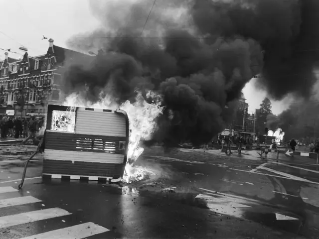 The image shows a black and white photo of a bus engulfed in flames on the side of a road, with a...