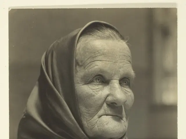 The image shows an old woman wearing a sweater and a headscarf. She has a wrinkled face and her...