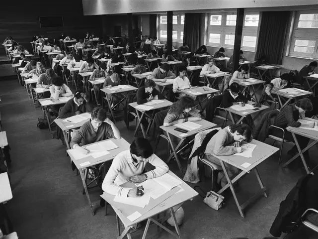 The image shows a black and white photo of a classroom full of students sitting at desks, writing...