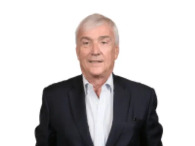 The image shows a man in a suit and tie standing in front of a white background, smiling. He...