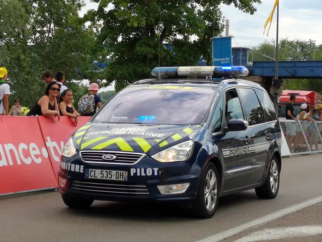 The image shows a police car driving down a street next to a crowd of people, some of whom are...