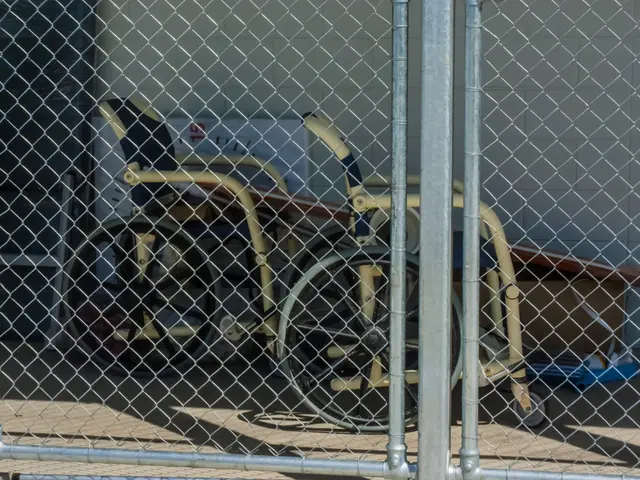 The image shows a wheelchair behind a chain link fence in front of a building. The wheelchair is...