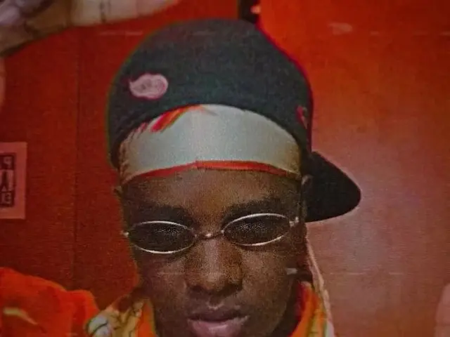 The image shows a man wearing glasses and a hat, with a chain around his neck, standing in front of...