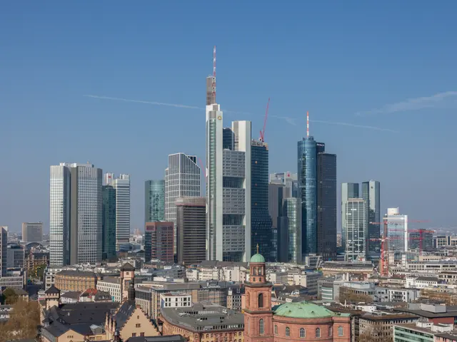 The image shows the Frankfurt skyline from the top of a hill, with a number of buildings and trees...
