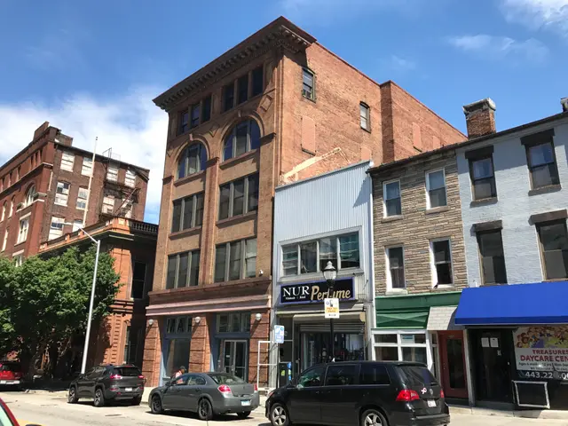 The image shows a city street with cars parked on the side of it, buildings with windows, trees,...