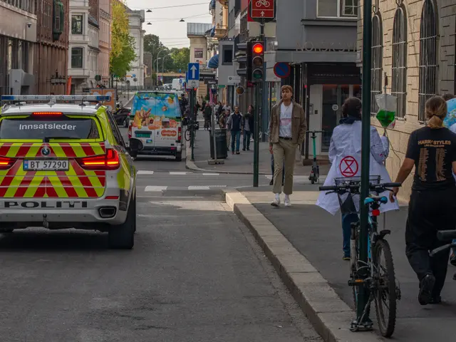 The image shows a police car driving down a street with a group of people walking down the sidewalk...
