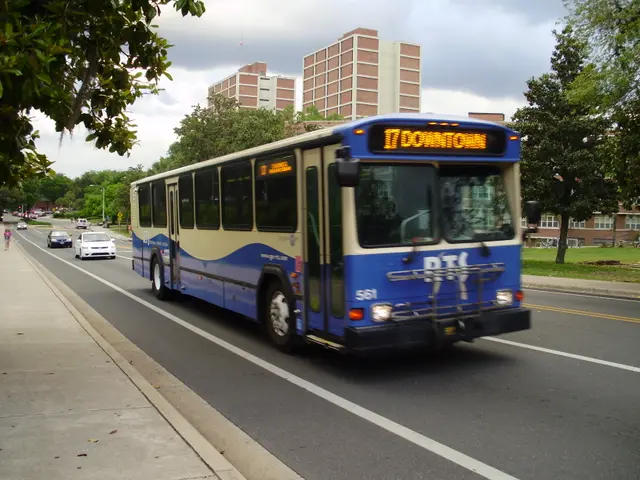 The image shows a blue and white shuttle bus driving down a street lined with tall buildings. On...