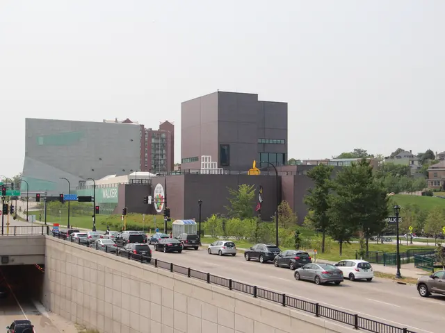 The image shows a city street with cars driving down it, a bridge with railings, light poles,...