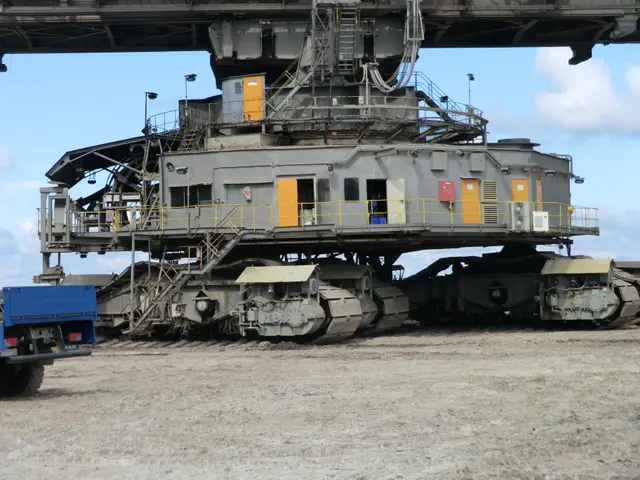 The image shows a large truck parked next to a large machine on top of a dirt field, with a bridge...