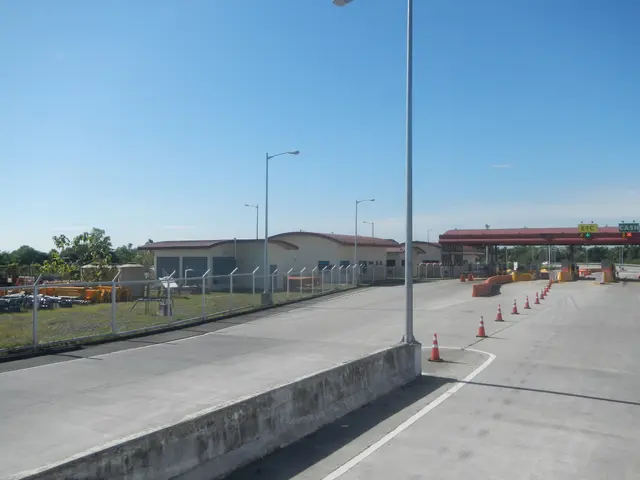 The image shows a road with traffic cones on the side of it, a metal fence, a group of buildings...