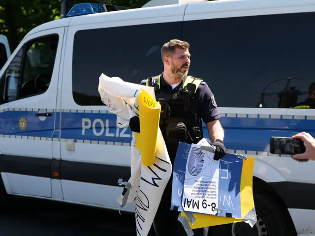 The image shows a police officer standing in front of a police van, holding a bunch of papers in...