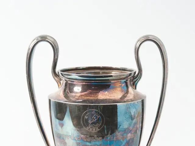 The image shows the UEFA Champions League Trophy, a steel cup with two handles and a blue and...