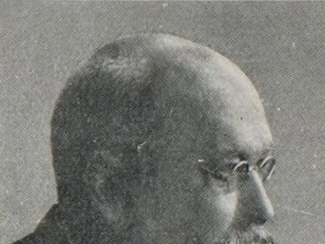 The image shows an old black and white photo of a man with a beard wearing a suit and spectacles....