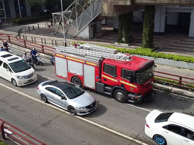 The image shows a fire truck driving down a street next to a car, with people riding motorbikes and...