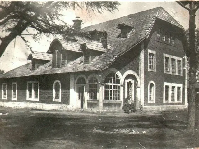 The image shows an old black and white photo of a building with windows, doors and a roof,...