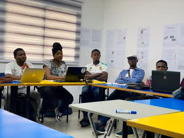 The image shows a group of people sitting around a table with laptops, bags, pens, and other...