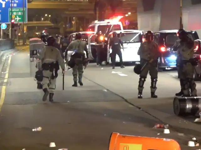 The image shows a group of police officers walking down a street at night. They are wearing helmets...