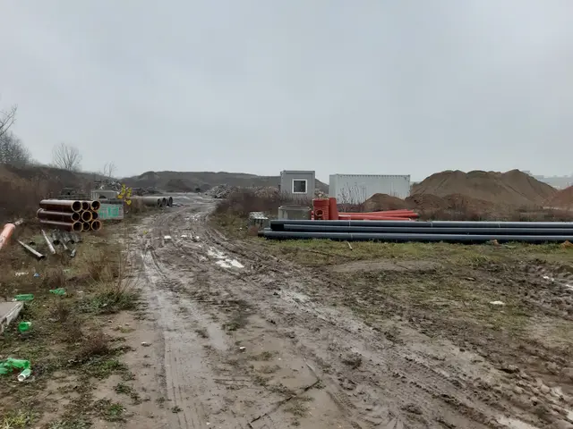 The image shows a dirt road with a lot of pipes on the side of it, surrounded by grass, plants,...
