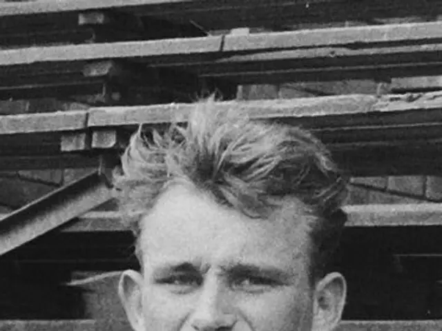 The image shows a black and white photo of a man wearing a jacket, standing in front of a pile of...