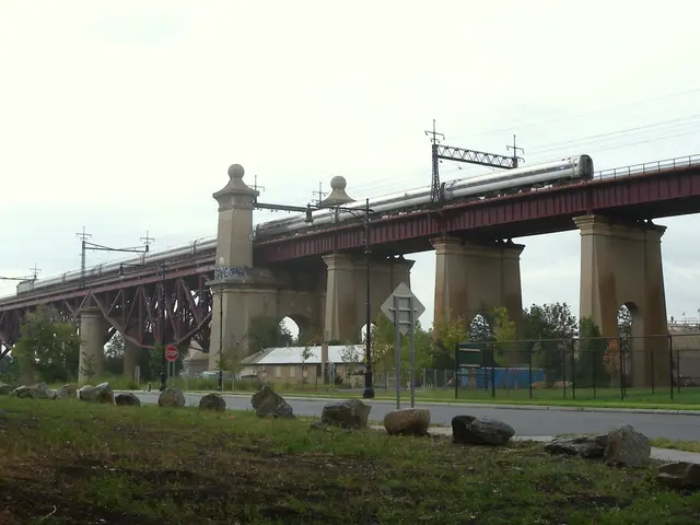 The image shows a train traveling over a bridge with pillars, electric poles with wires, sign...