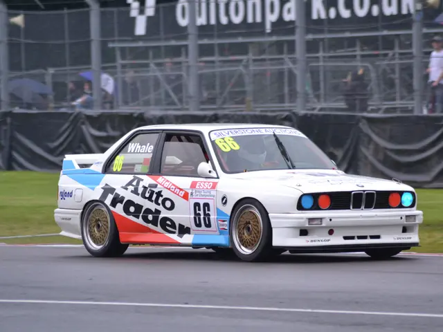 The image shows a white car driving down a race track with text and numbers on it, surrounded by...