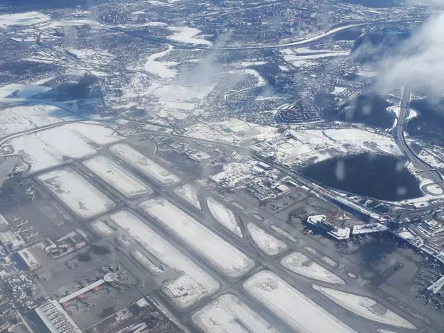The image shows an aerial view of an airport with snow on the ground, buildings, roads, bridges,...