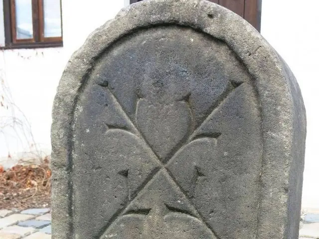 The image shows a stone headstone with a cross on it in front of a building with windows and a...
