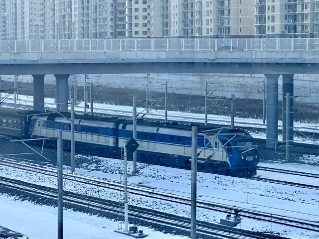 The image shows a blue and white train traveling down train tracks next to tall buildings, with...