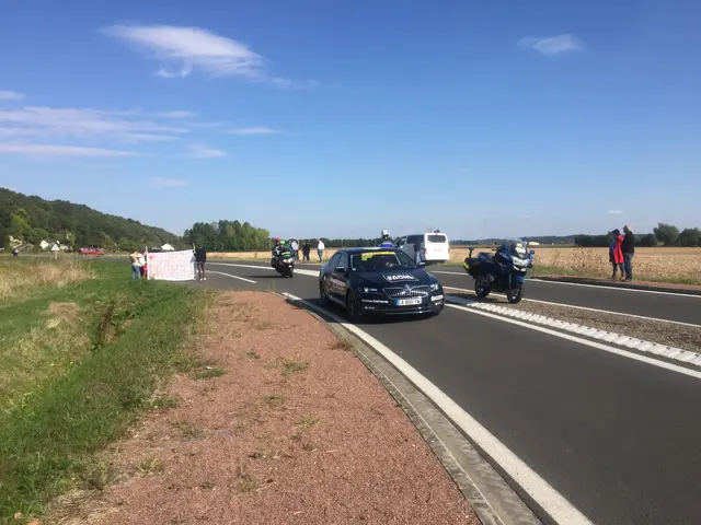 The image shows a group of people standing on the side of a road next to a car, with a banner with...