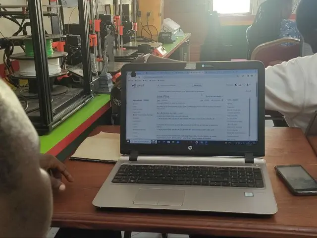 The image shows a group of people sitting at a table with laptops, mobile phones, books, papers,...