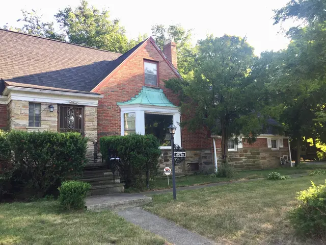 The image shows a brick house with a green awning in front of it, surrounded by plants, grass, a...