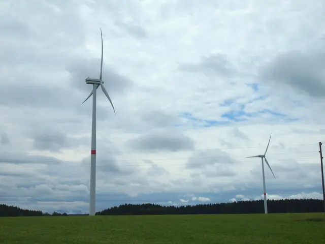 The image shows three wind turbines in a green field with trees in the background and clouds in the...