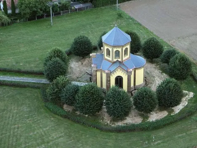 The image shows an aerial view of a small church in the middle of a field surrounded by trees,...