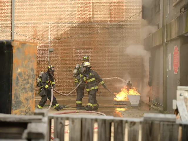 The image shows a group of firefighters wearing helmets and holding pipes in their hands, working...