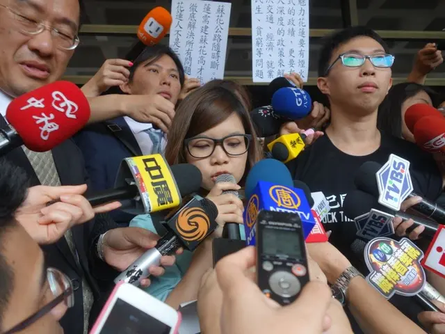 The image shows a group of people standing in front of a crowd of reporters, some of whom are...