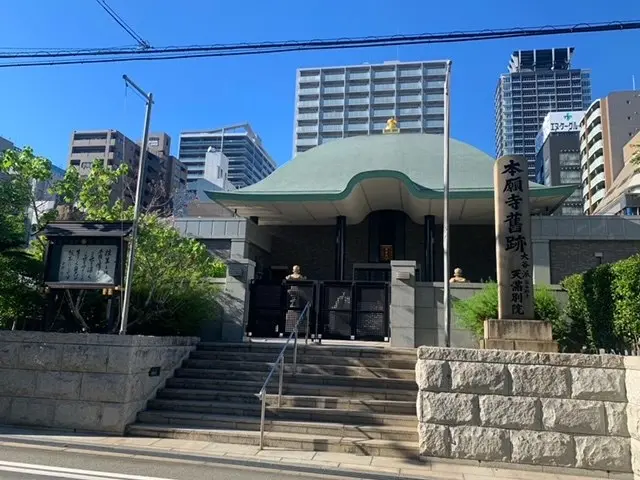 The image shows a large building with a green roof in the middle of a city, surrounded by stairs,...