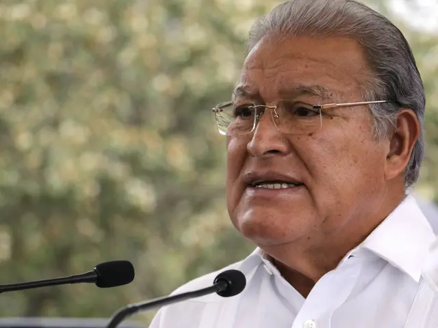 The image shows a man in a white shirt and glasses speaking into a microphone in front of a blurred...