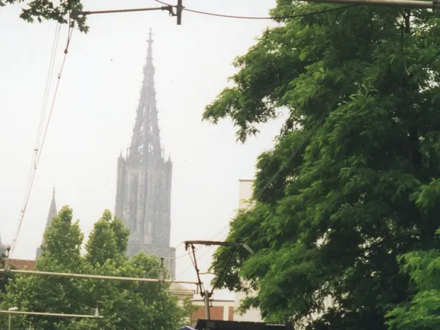The image shows a red and white train traveling down tracks next to a tall building, with a person...