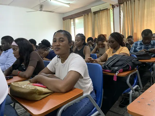 The image shows a group of people sitting at desks in a classroom, with bags and other objects on...