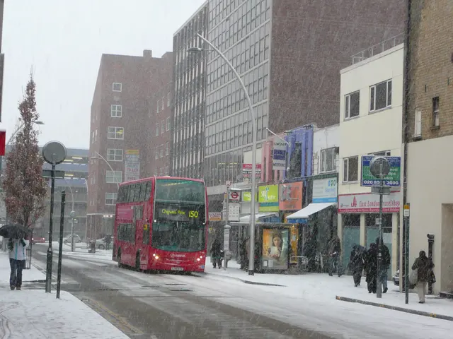 The image shows a red double decker bus driving down a snowy street, surrounded by tall buildings....