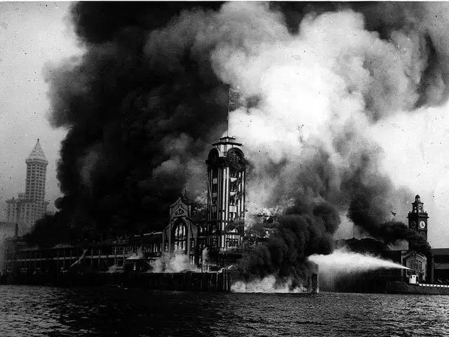 The image shows a black and white photo of a large building engulfed in flames, with smoke...