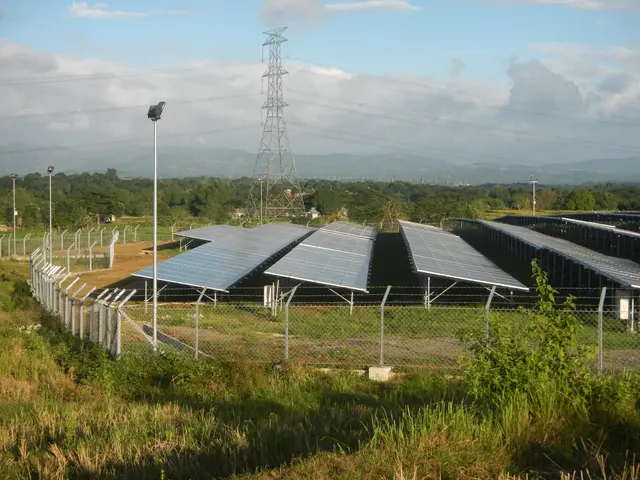 The image shows a field of solar panels with a fence in the foreground, surrounded by grass,...