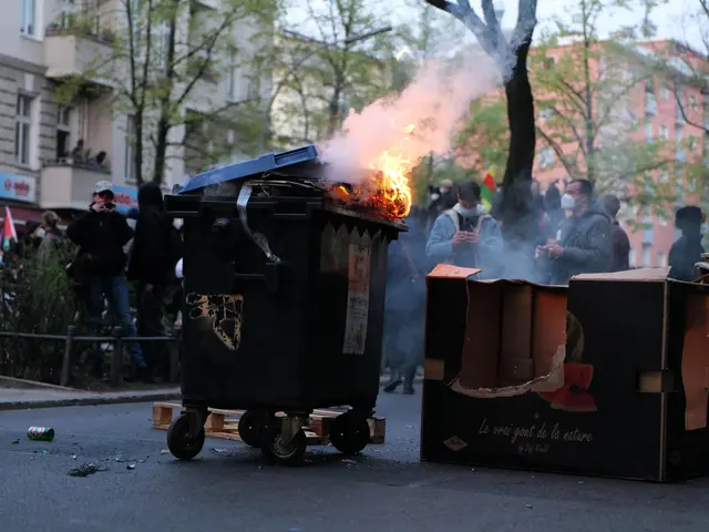 The image shows a group of people standing around a trash can with fire coming out of it,...