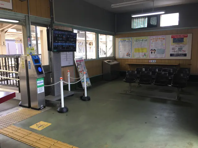 The image shows a train station with a lot of chairs and a counter, as well as machines, poles with...